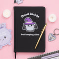 Black notebook with a cute ghost design and text on a pink background with decorative items.