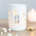 White candle holder with leaf cutouts on a wooden surface with a lit candle in the background.