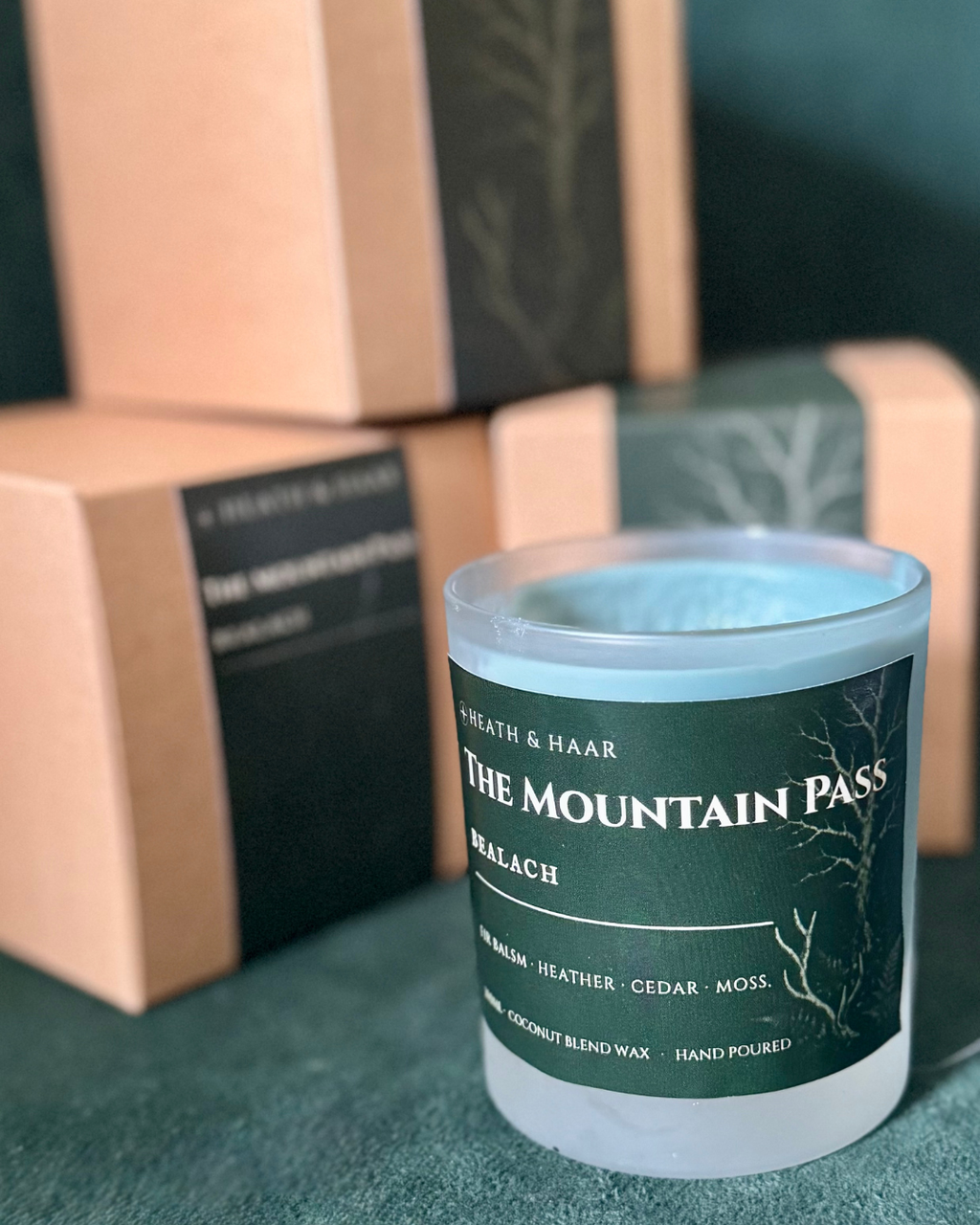 A frosted glass candle jar labeled 'THE MOUNTAIN PASS' ' sits in the foreground, with matching kraft paper gift boxes blurred in the background on a dark green surface.