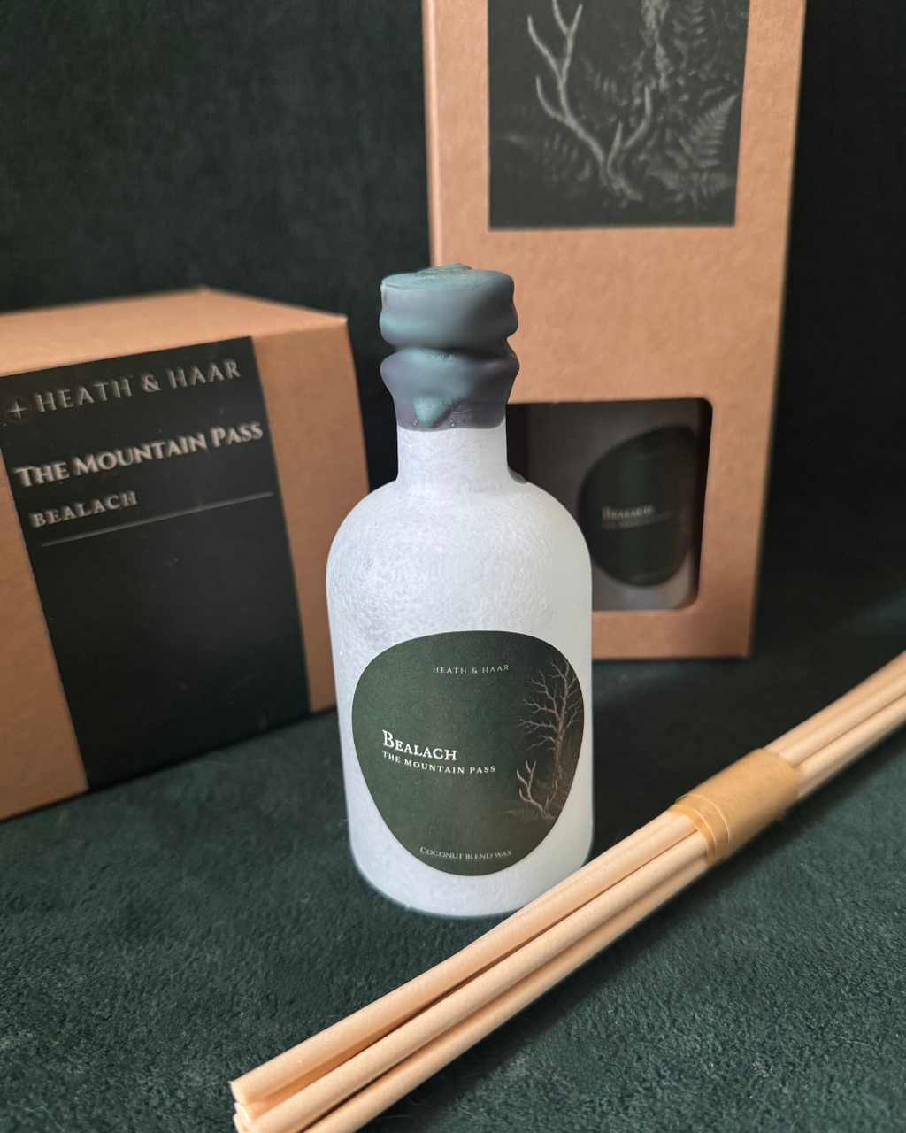 A frosted glass bottle with a dark green label reading THE MOUNTAIN PASS' and 'HEATH & HAAR', shown with a matching brown gift box and a bundle of wooden reeds on a dark green surface.