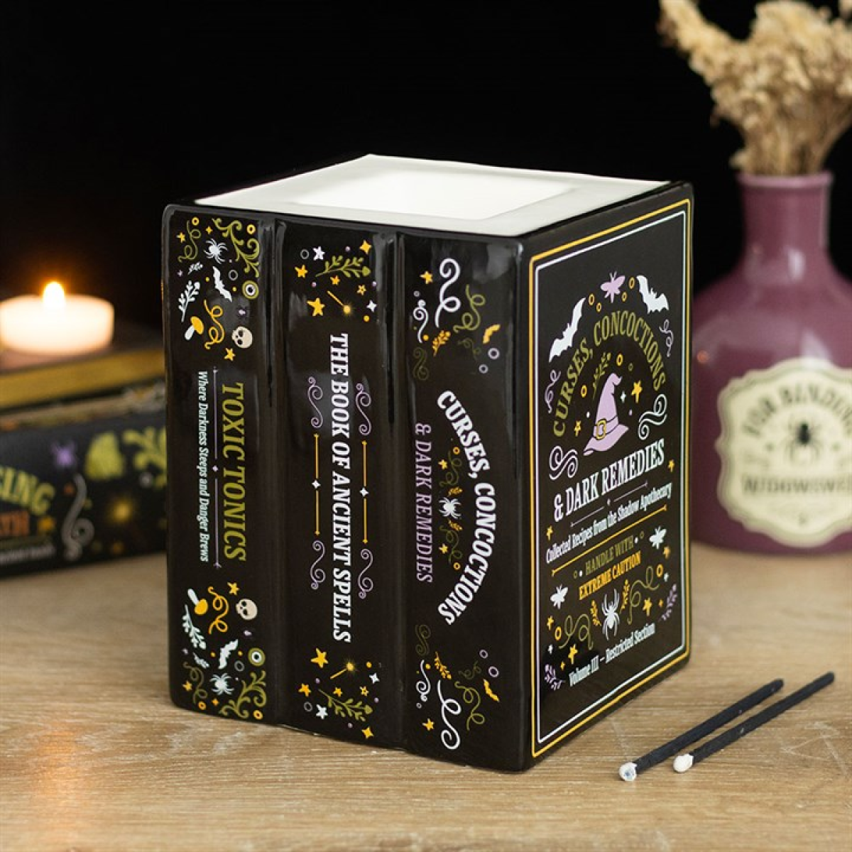 Three black books with decorative covers on a wooden surface, with a candle and vase in the background.