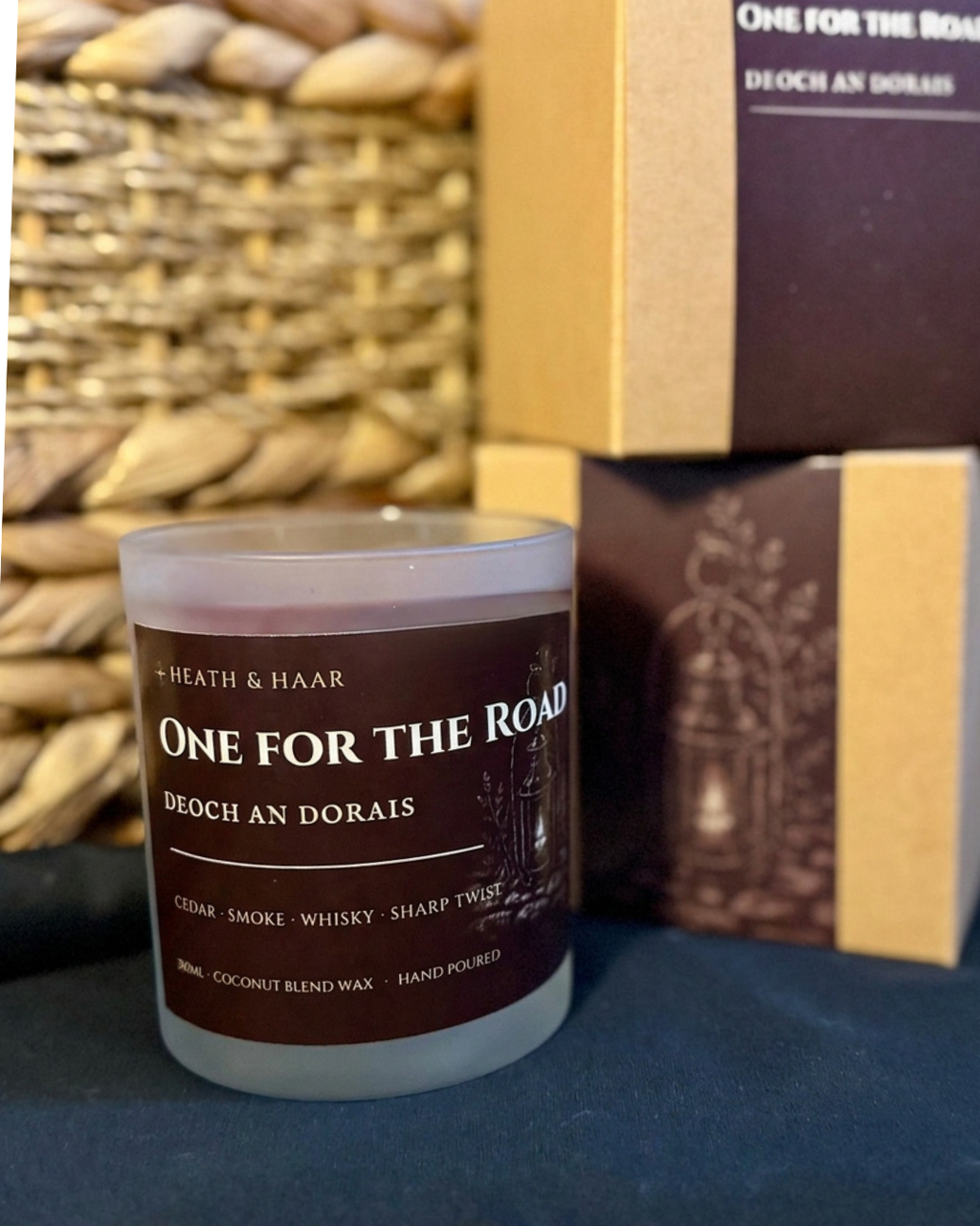 A jar candle labeled “One For The Road” by Heath & Haar sits in front of stacked product boxes on a dark surface, with a woven basket blurred in the background.