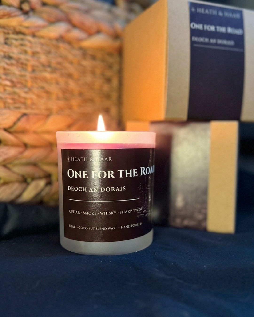 A lit Heath & Haroar candle in a frosted glass jar with a dark brown label reading 'ONE FOR THE ROMANTIC', set on a dark blue surface with a woven basket and product boxes in the background.