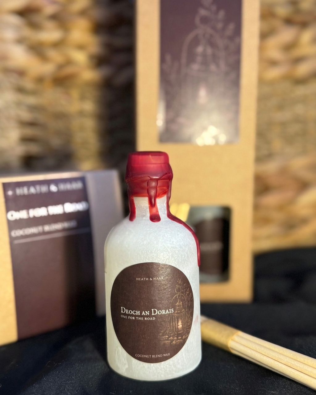 A small frosted glass bottle candle with a red wax seal and a brown circular label reading 'Droce as Dorari' by Heath & Rams, shown with its packaging box and wooden wick accessories on a dark surface.