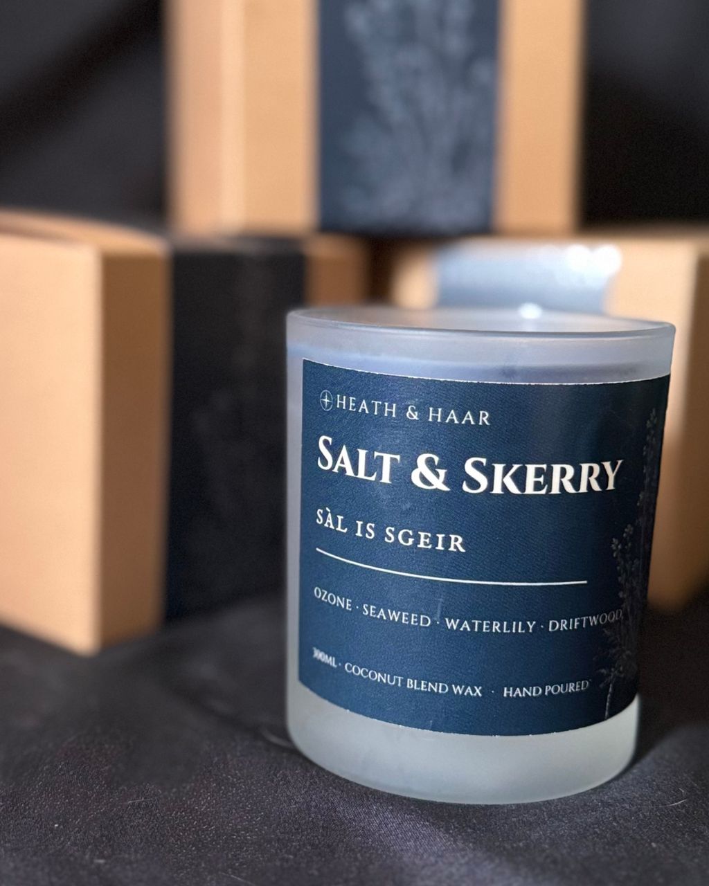 A frosted glass jar candle with a dark navy blue label reading 'HEATH & HAAR SALT & SKERRY', photographed on a dark surface with blurred kraft paper boxes in the background.