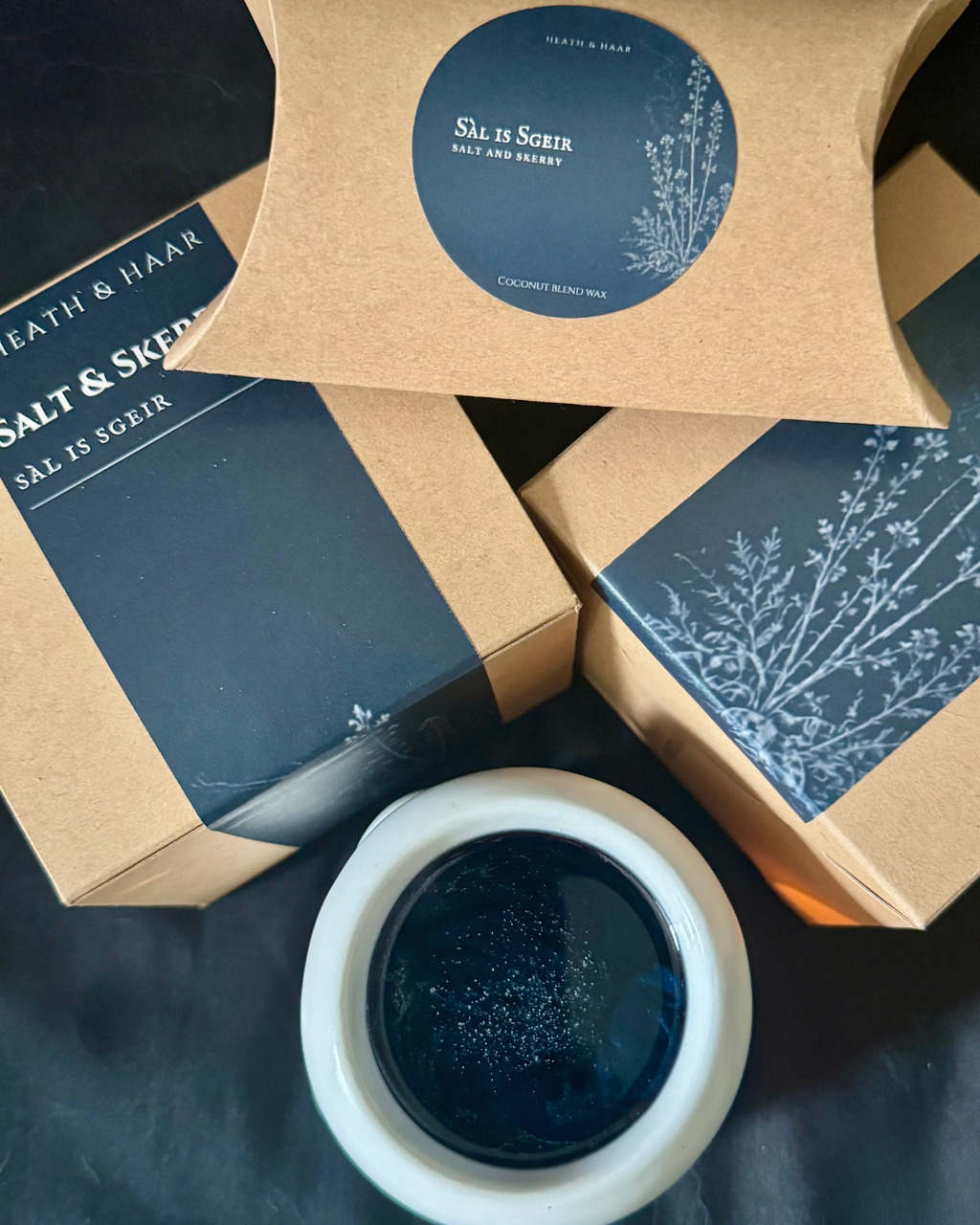 Three brown kraft paper boxes of 'Salt & Skerryr' coconut wax melt are arranged on a dark surface, with one box showing a navy blue label and a white ceramic bowl containing the dark blue wax product in the foreground.