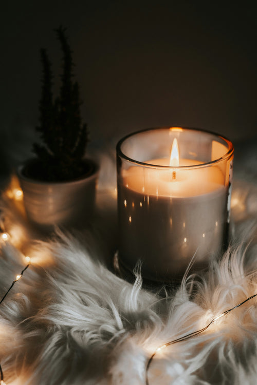 Candle burning on a fur surface with decorative lights and a plant in the background